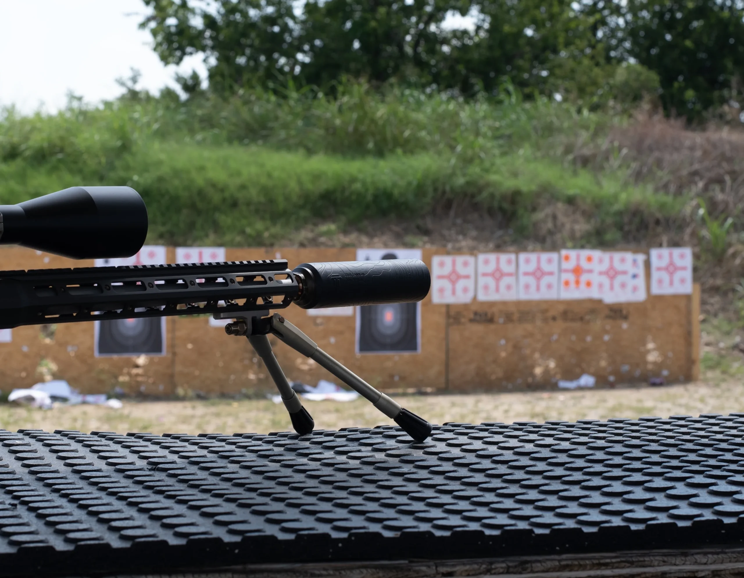 Suppressed precision rifle on a bipod at an outdoor range with paper targets on the berm